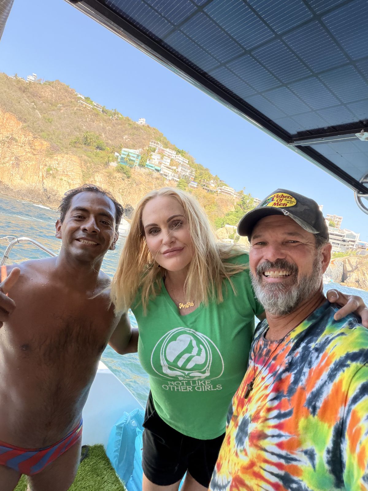 A cliff diver in red and blue trunks giving a thumbs up, with Meghan and Steve, on the back of Some Tuesday. Cliffs and houses behind them.