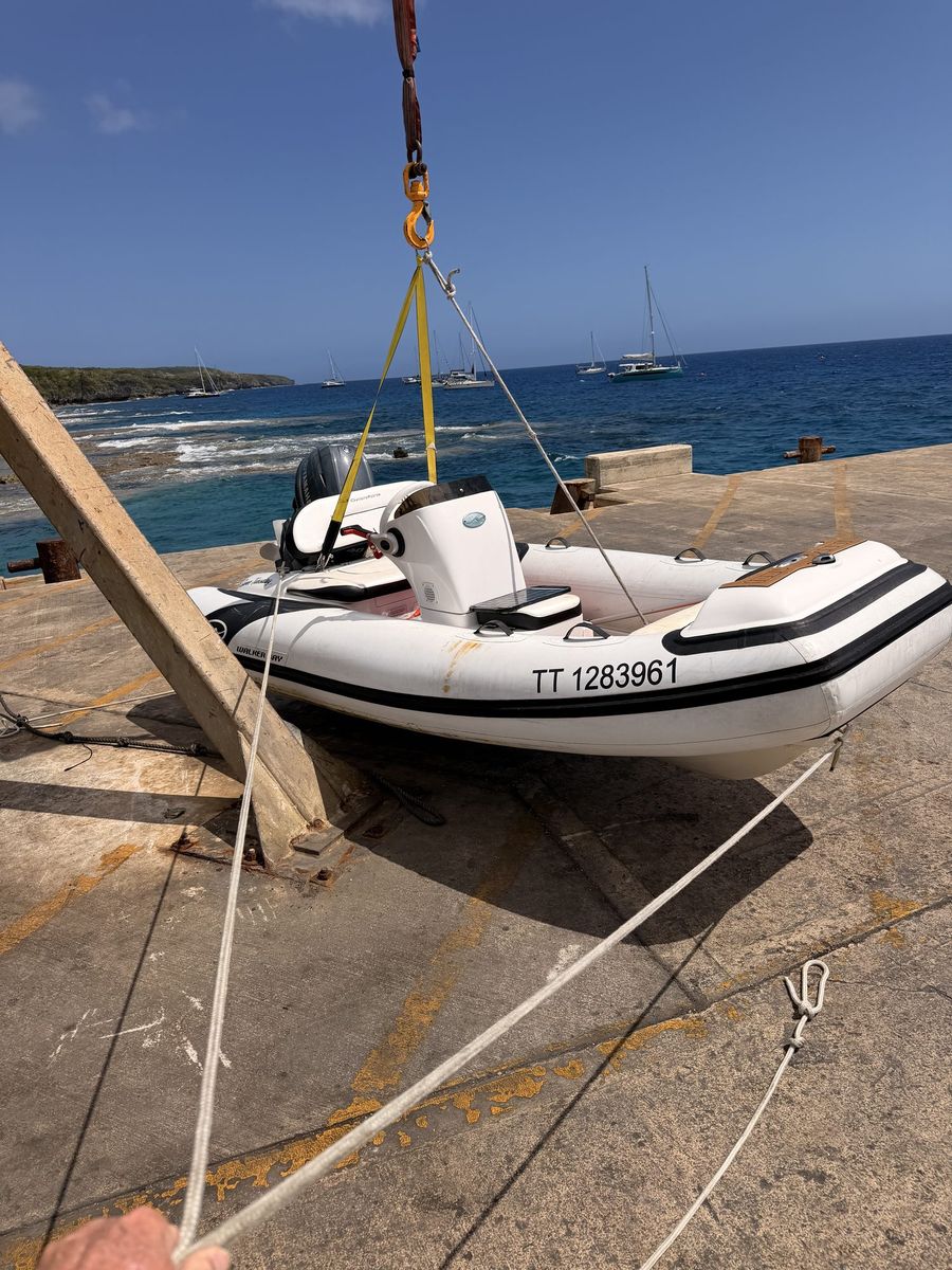 A white dinghy hanging in yellow crane straps above a concrete ramp, with a hand holding a line and sailboats offshore