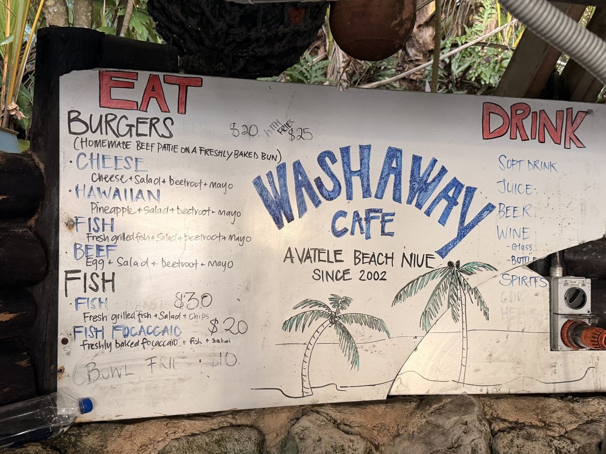A painted menu board reading Washaway Cafe, Avatele Beach, Niue, with burger and fish prices
